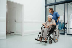 Portrait of nurse pushing senior patient in a wheelchair across hospital corridor, hall. Emotional support for elderly woman.